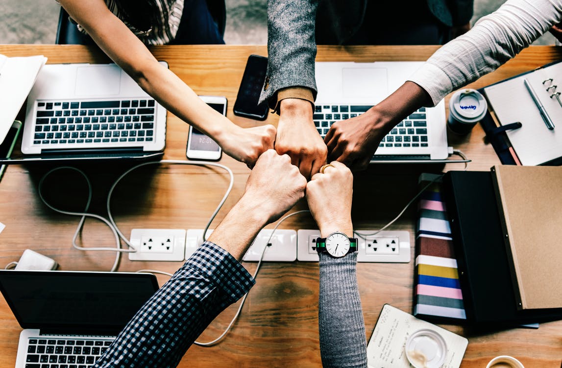 Five people fist-bumping over a desk with laptops and folders on it